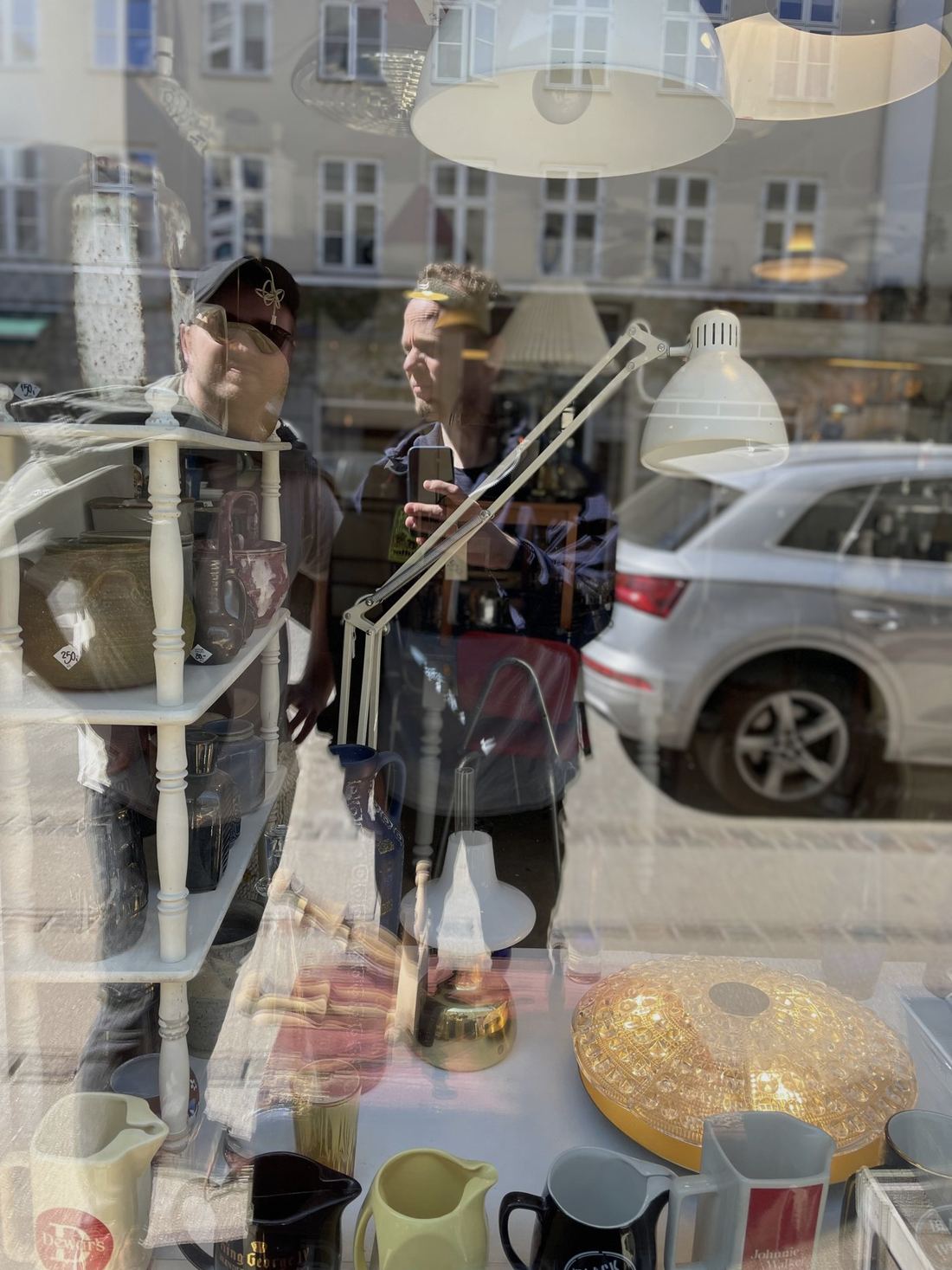 Elliott and Jacob reflected in a shop window, among lamps and jugs, the street behind them