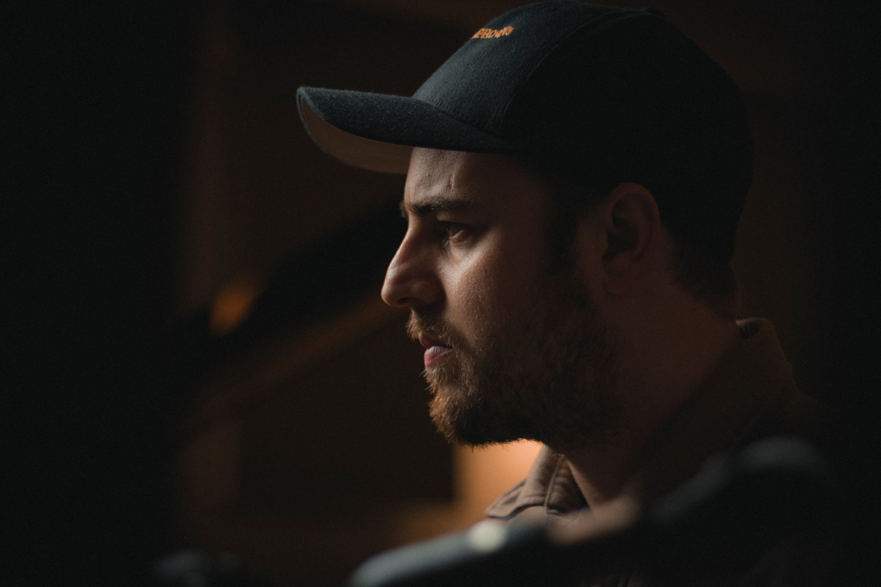 Close profile of Elliott in a black cap, lit warmly from the side against a dark background